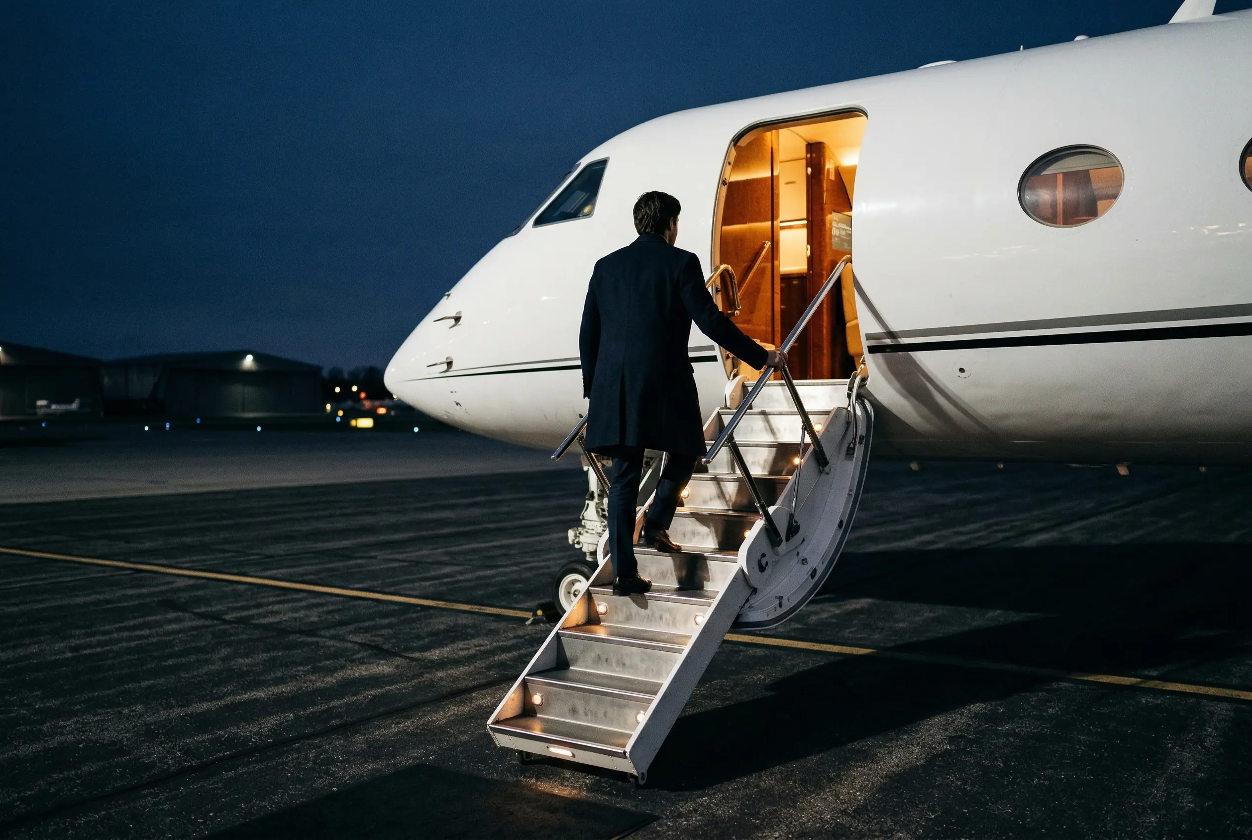 Executive boarding a private jet via airstairs at dusk — warm cabin light illuminating the entrance on a quiet New York ramp